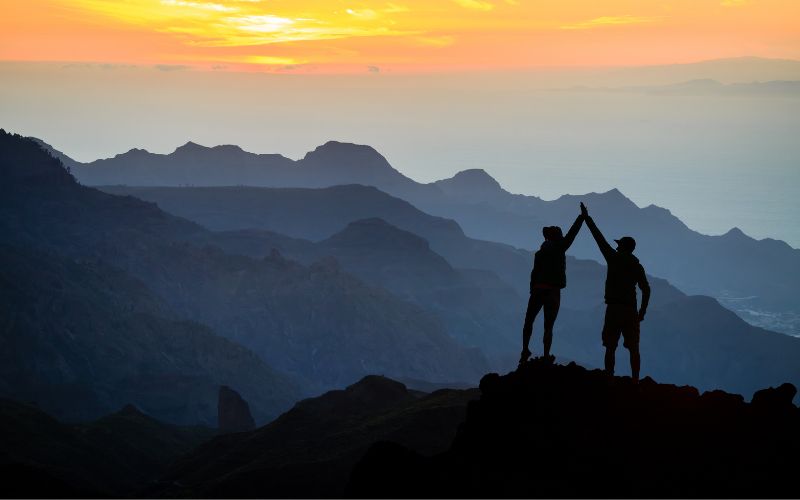 silhouette of two people high-fiving at the top of a peak in the mountains