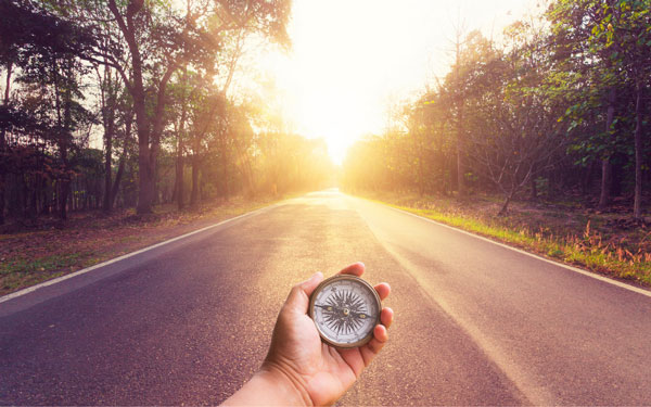 a hand holding a compass with a road leading to sunrise ahead