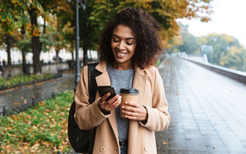 woman walking outside smiling while looking at phone and holding a coffee cup
