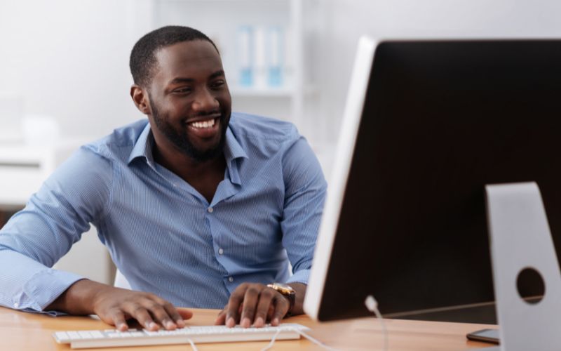 man with blue button down shirt smiling from behind a computer monitor while typing